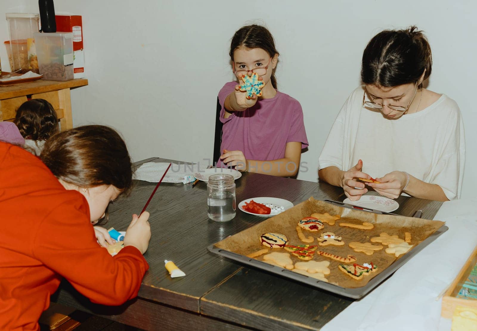 Three beautiful Caucasian brunettes are sitting at the table and two are enthusiastically decorating baked cookies with mastic and chocolate, and the third is showing her cookies, close-up side view. Step by step instructions. Step 26