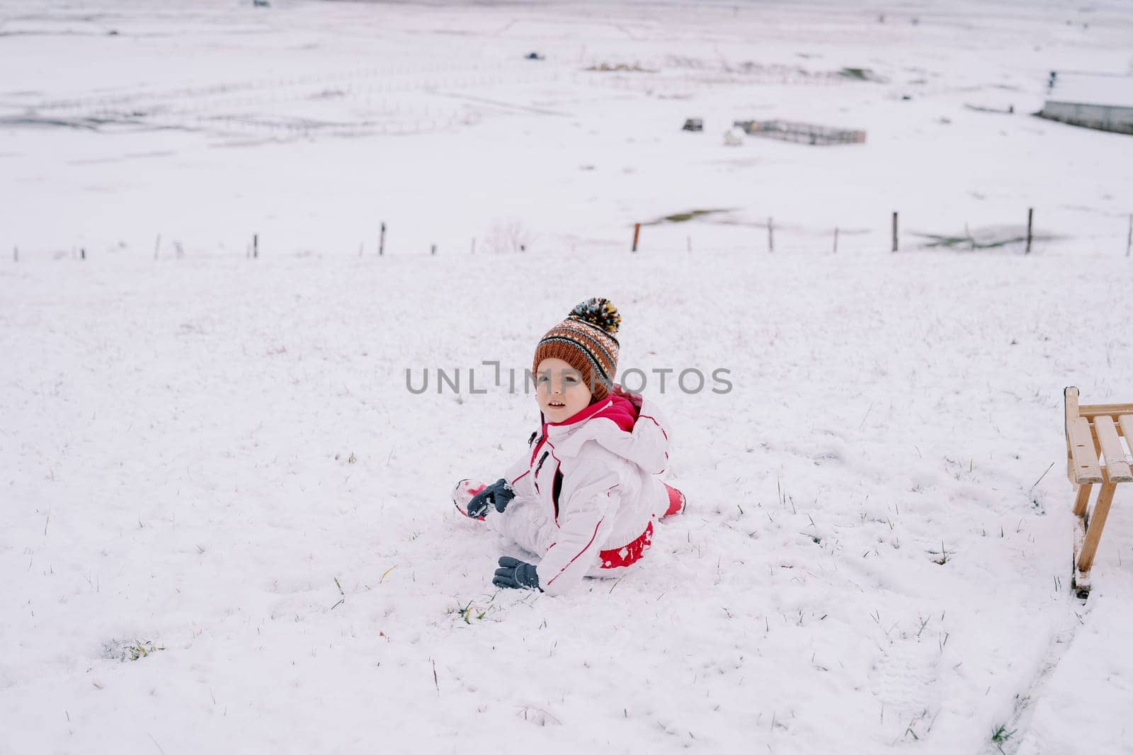 Little girl sits half-turned on the snow near a sled. High quality photo