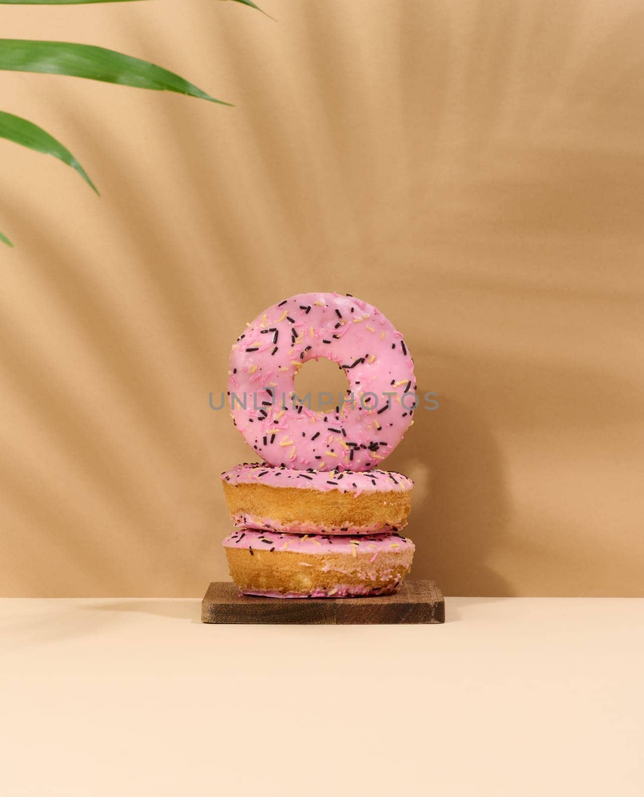 Donut covered with pink glaze and sprinkled with multi-colored sprinkles, brown background with a shadow from a palm leaf