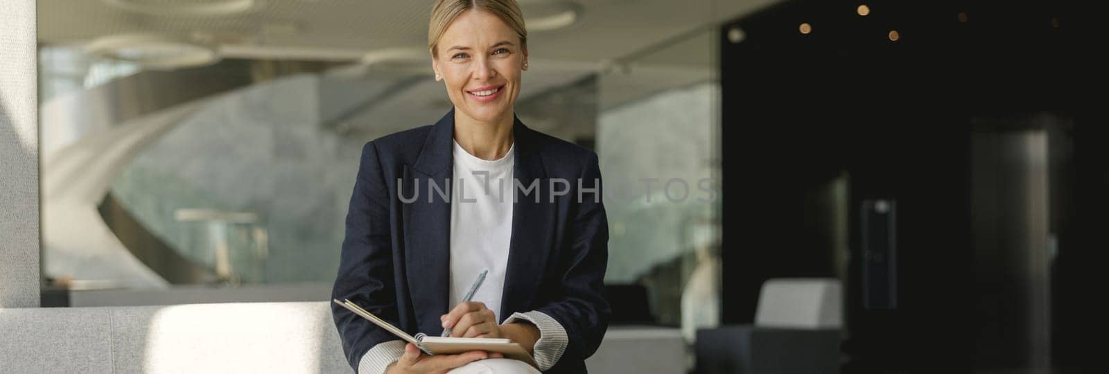 Pretty businesswoman making notes in note pad sitting on modern office background by Yaroslav_astakhov