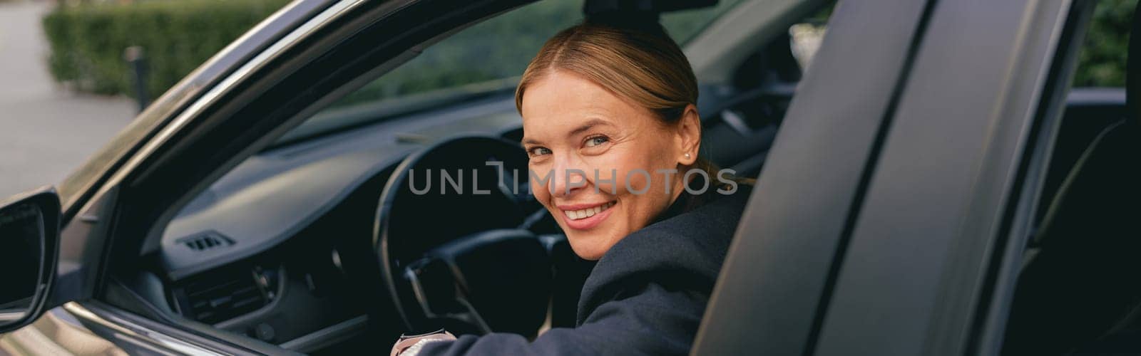 Female driver in suit is riding behind steering wheel of car and looks camera. High quality photo