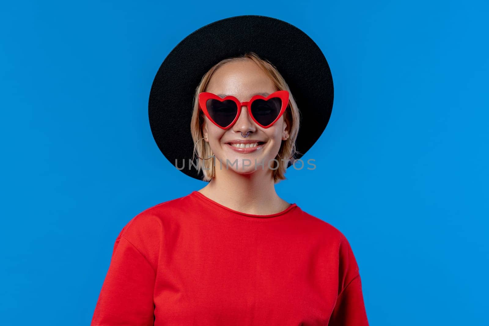 Hipster woman in black hat and heart-shaped eyewear on blue background. Beautiful lady with european appearance looking at camera. High quality photo
