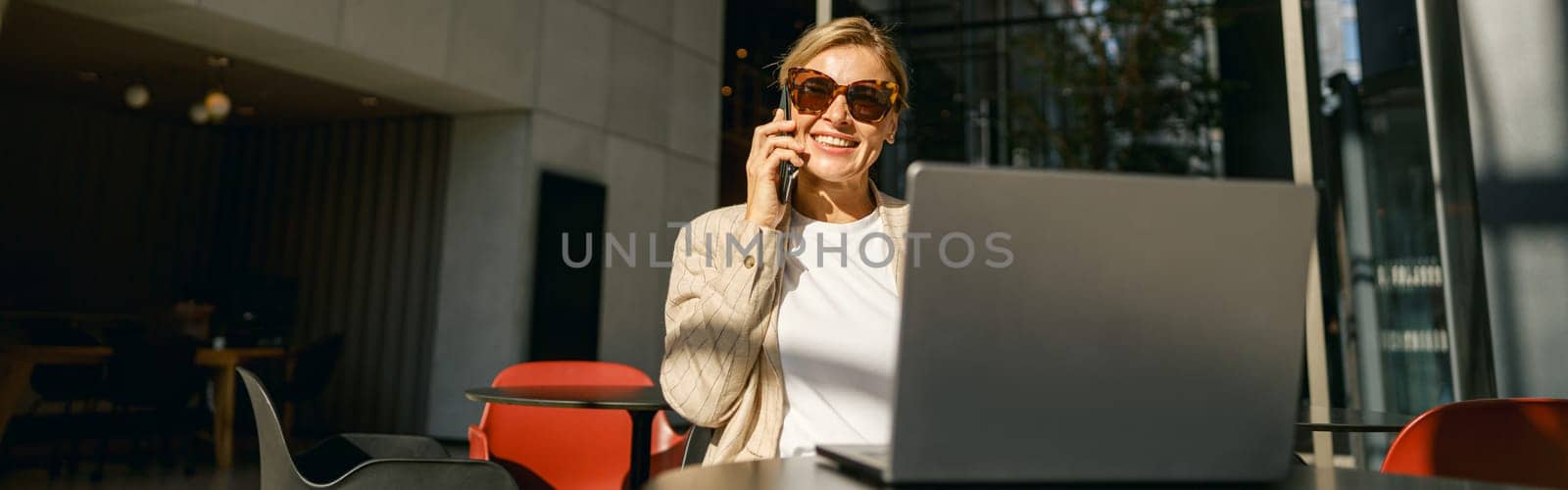 Woman freelancer talking on phone with client while working on laptop in modern coworking by Yaroslav_astakhov