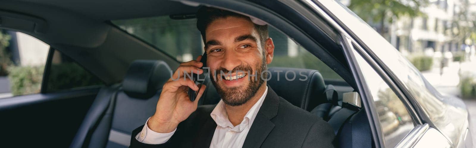 Smiling businessman talking phone with client while sitting on car backseats by Yaroslav_astakhov