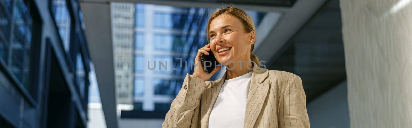 Smiling business woman holding laptop and talking phone standing on modern building background by Yaroslav_astakhov