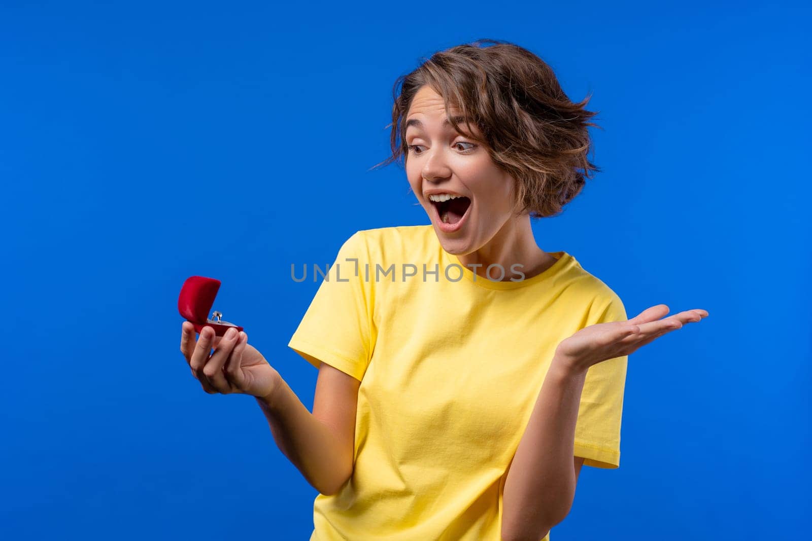 Pretty woman got jewelry box with proposal diamond ring on blue wall background. Surprised lady smiling, she is happy to be fiancee, proposition for marriage. High quality photo