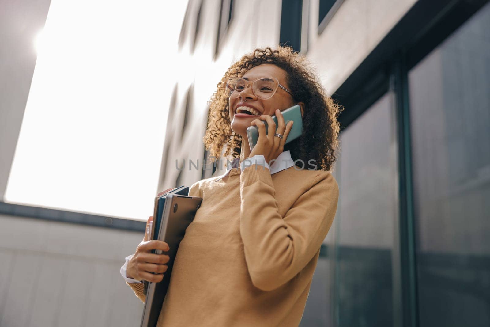 Smiling woman manager is talking phone while standing with laptop on modern building background