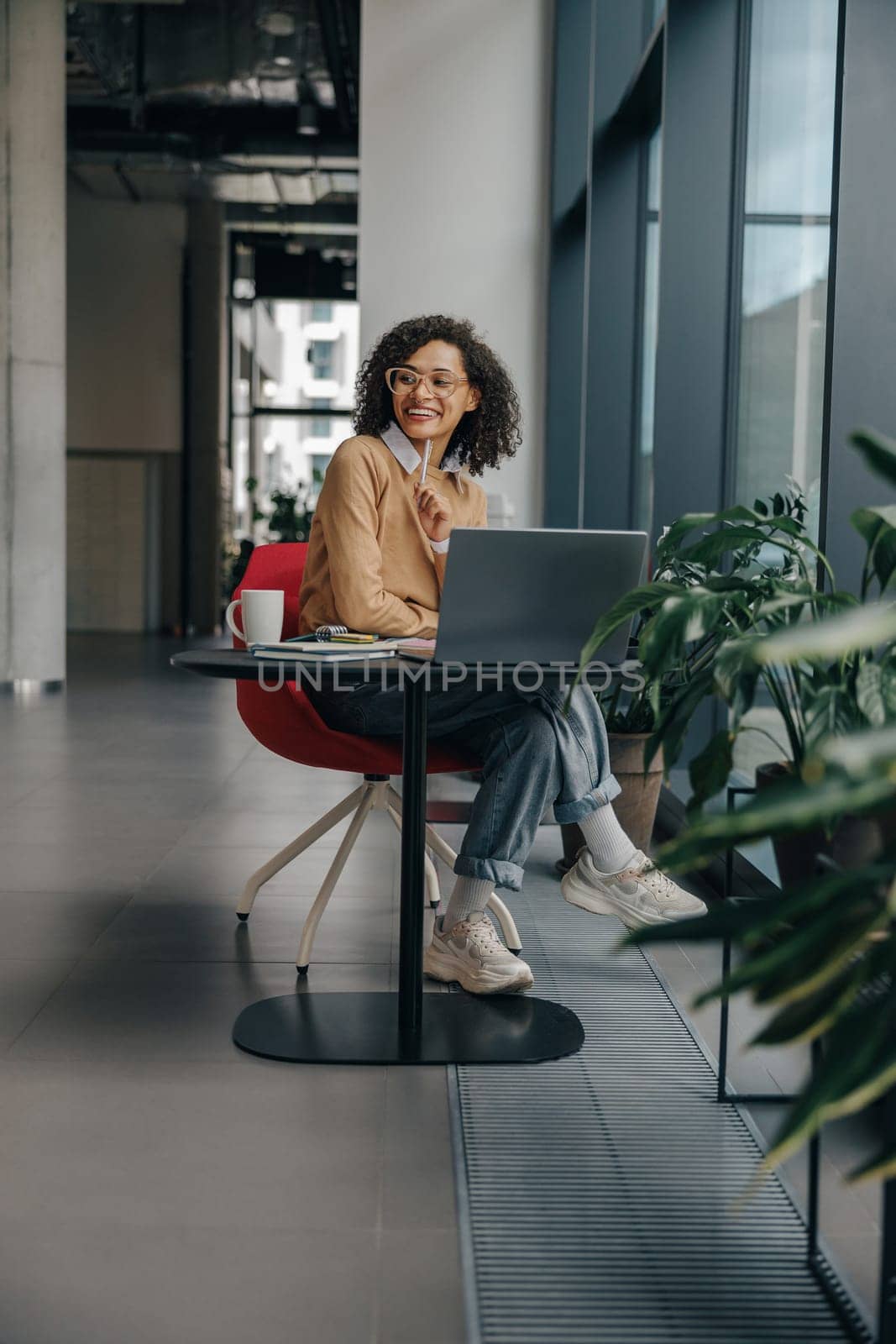 Smiling female manager in eyeglasses working on laptop while sitting in modern office near window by Yaroslav_astakhov