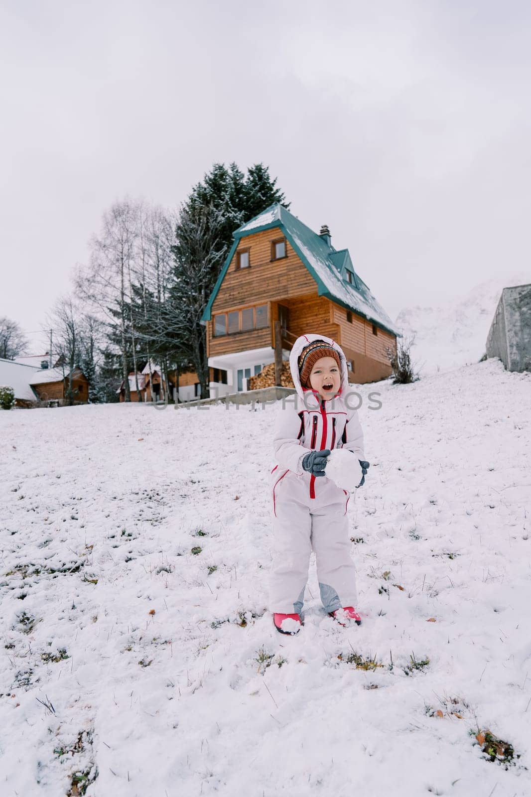 Little laughing girl with a snowball in her hands stands on a snowy slope near a wooden house. High quality photo