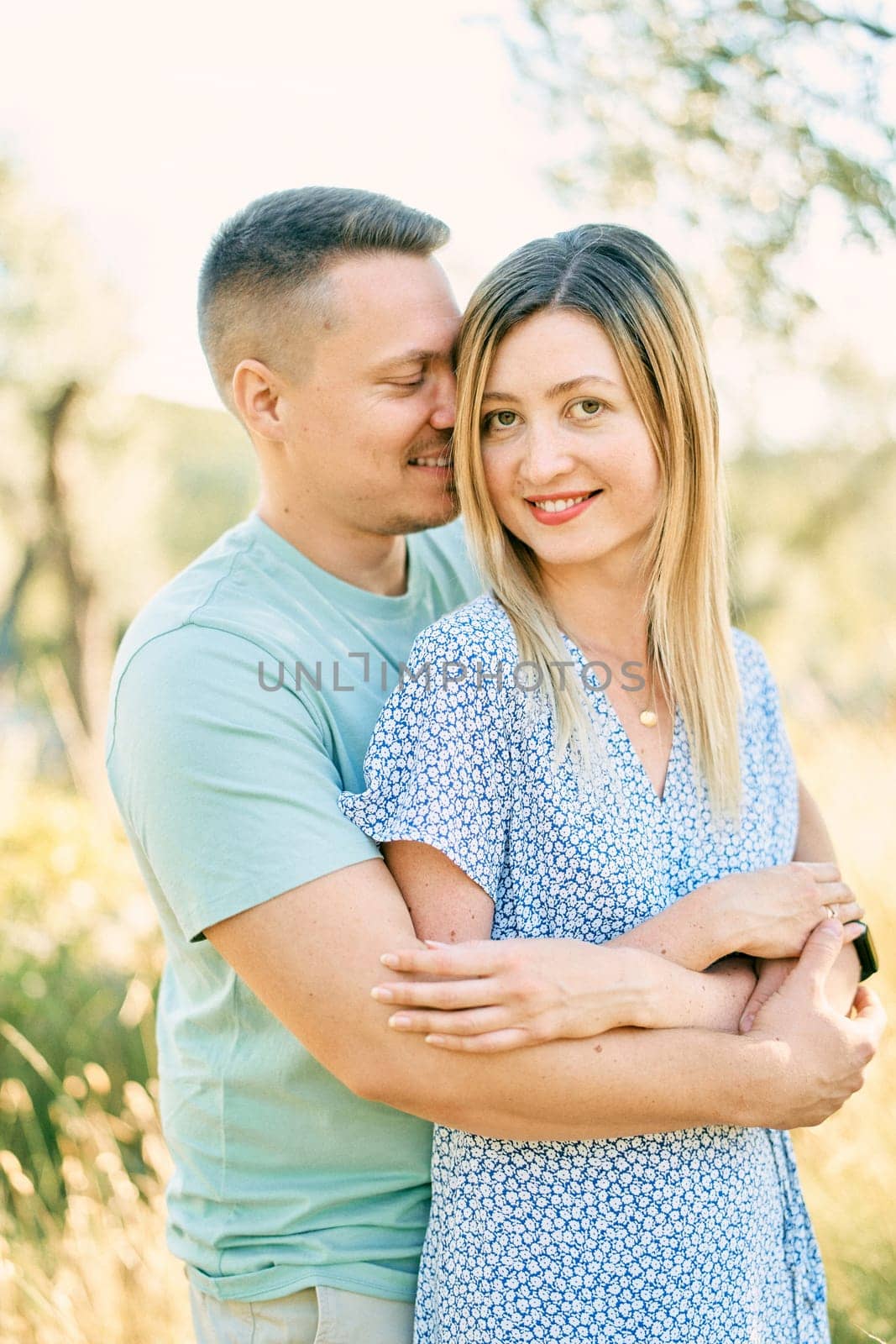 Guy hugs smiling girl from behind, touching her cheek with his nose while standing in the garden. High quality photo