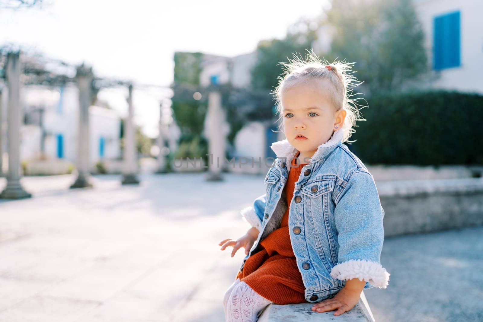 Little girl sits on a stone fence in the yard and looks away. High quality photo