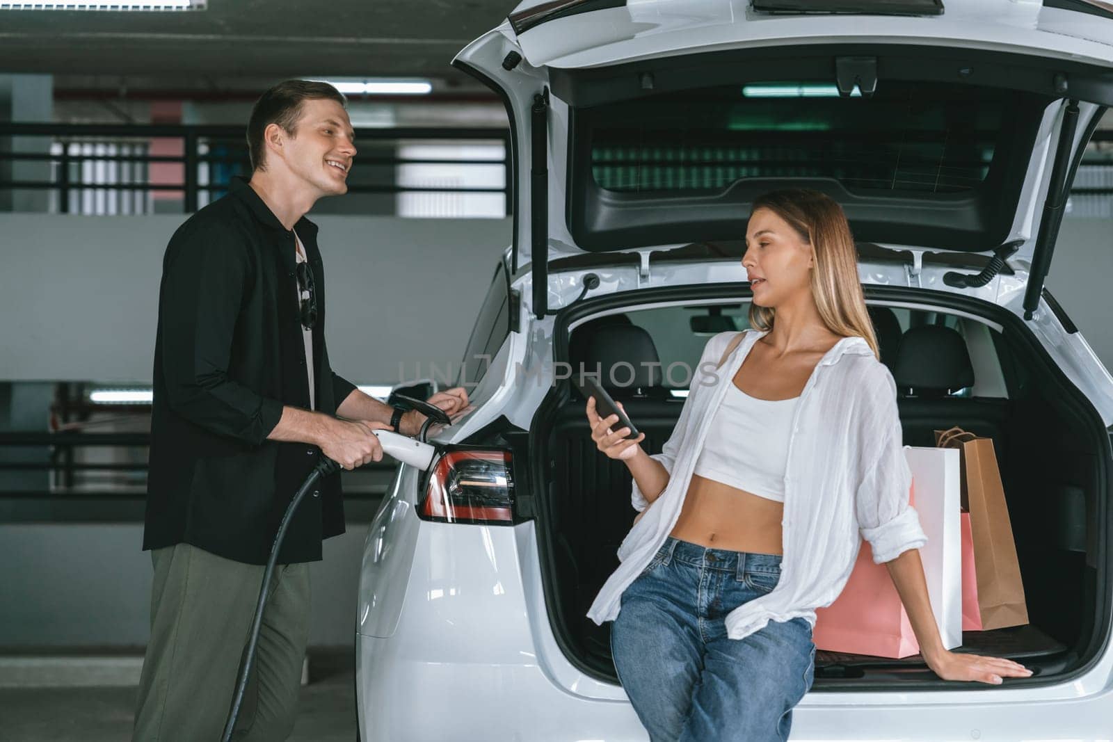 Young couple travel with EV electric car to shopping center parking lot charging in downtown city showing urban sustainability lifestyle by green clean rechargeable energy of electric vehicle innards