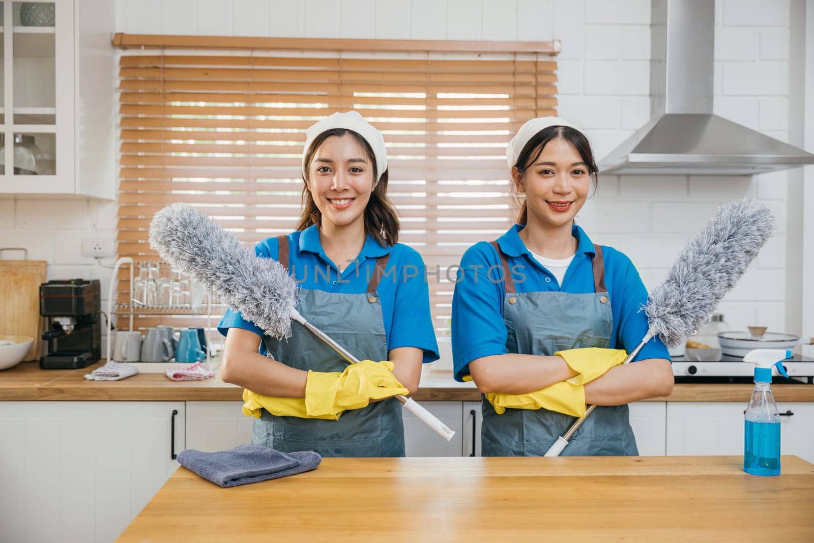 Portrait of two Asian housekeepers on kitchen counter with duster foggy spray and rag. Their teamwork effective housework and hygiene. Clean portrait two uniform maid working smiling employee.