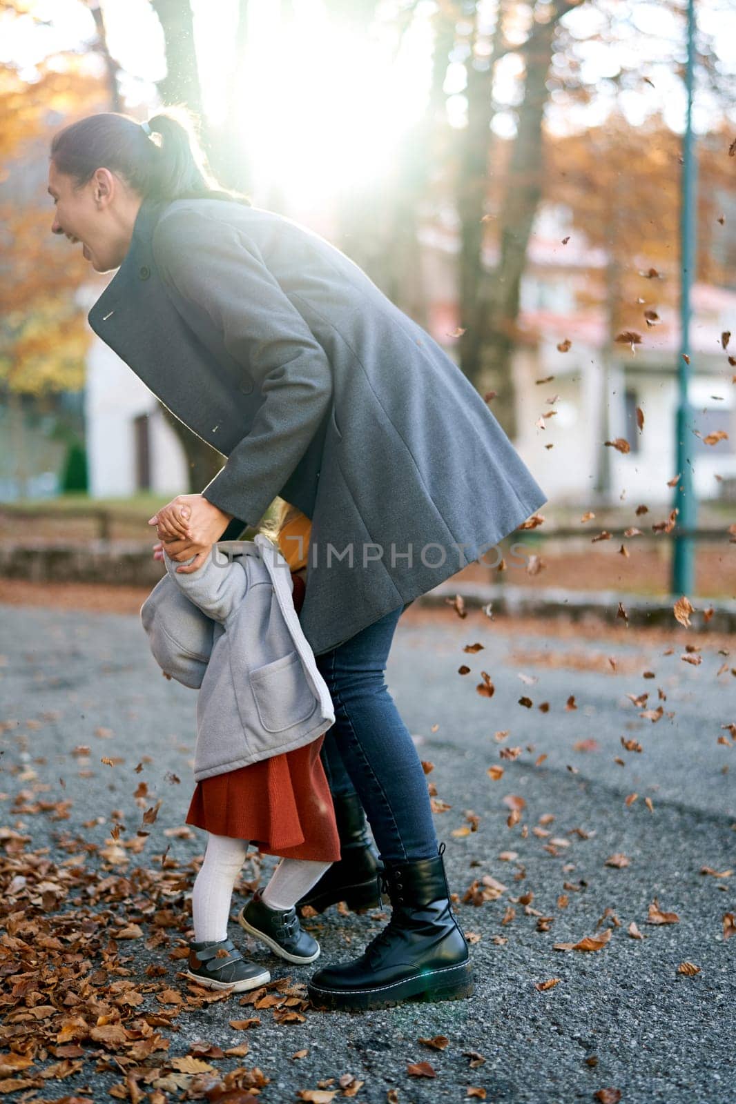 Smiling mom holding hands a little girl hiding her face in her lap while standing under falling leaves. High quality photo