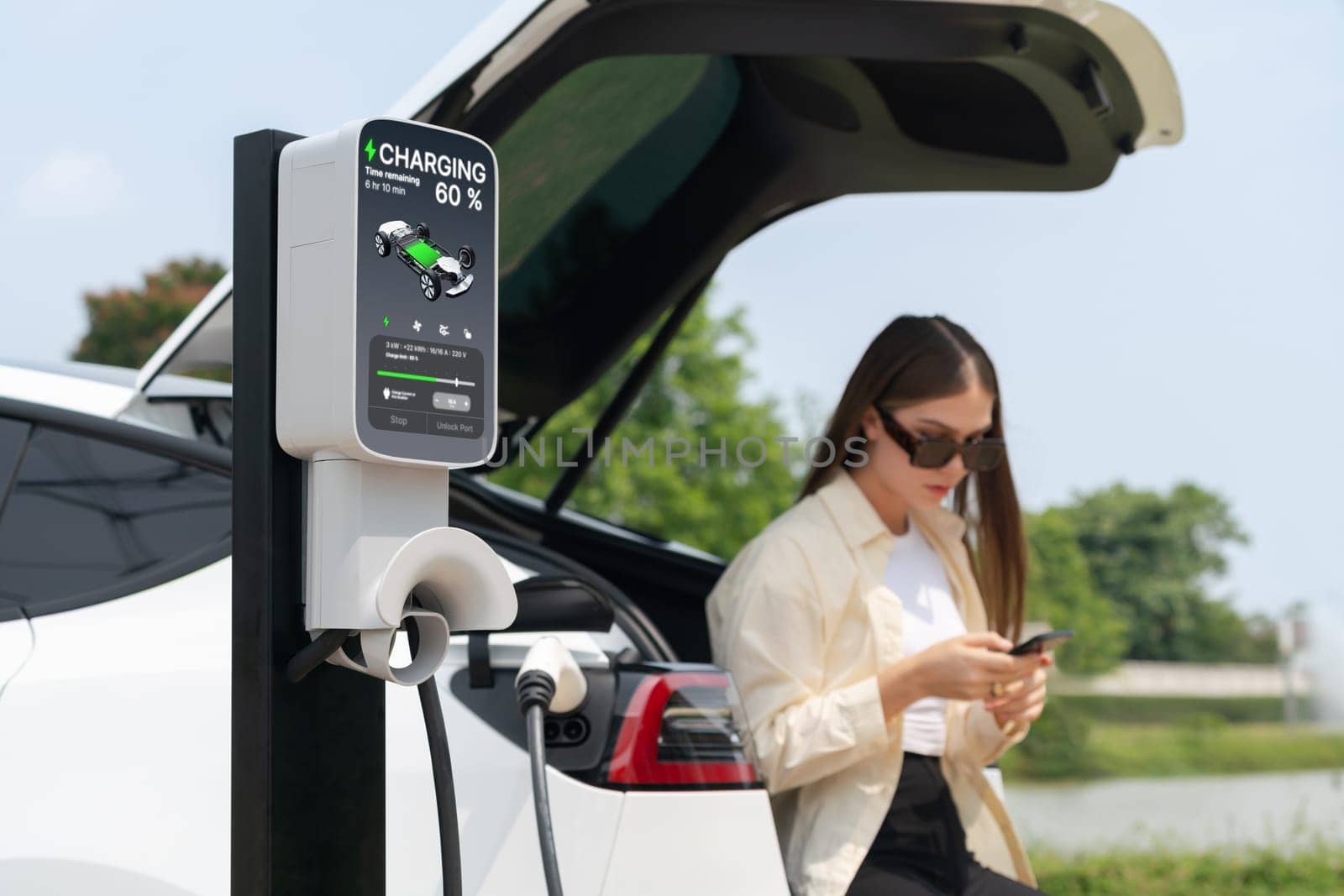 Young woman use smartphone to pay for electricity at public EV car charging station green city park. Modern environmental and sustainable urban lifestyle with EV vehicle. Expedient