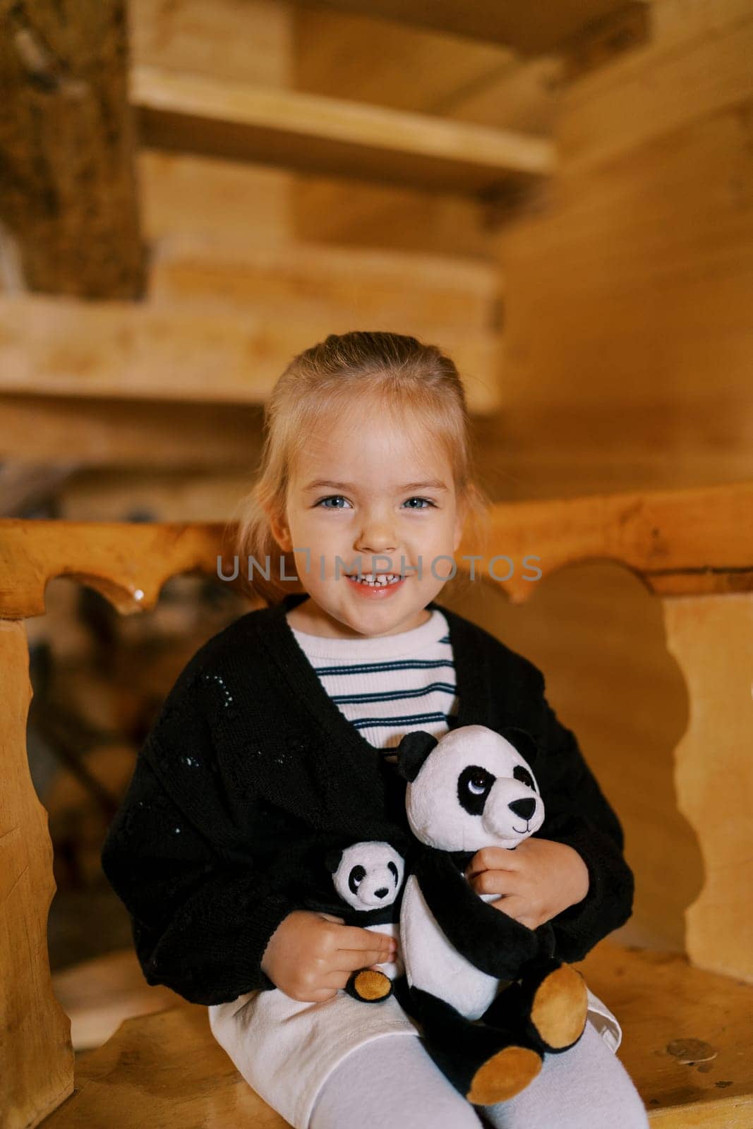 Little smiling girl sitting with soft toy pandas on a wooden chair. High quality photo
