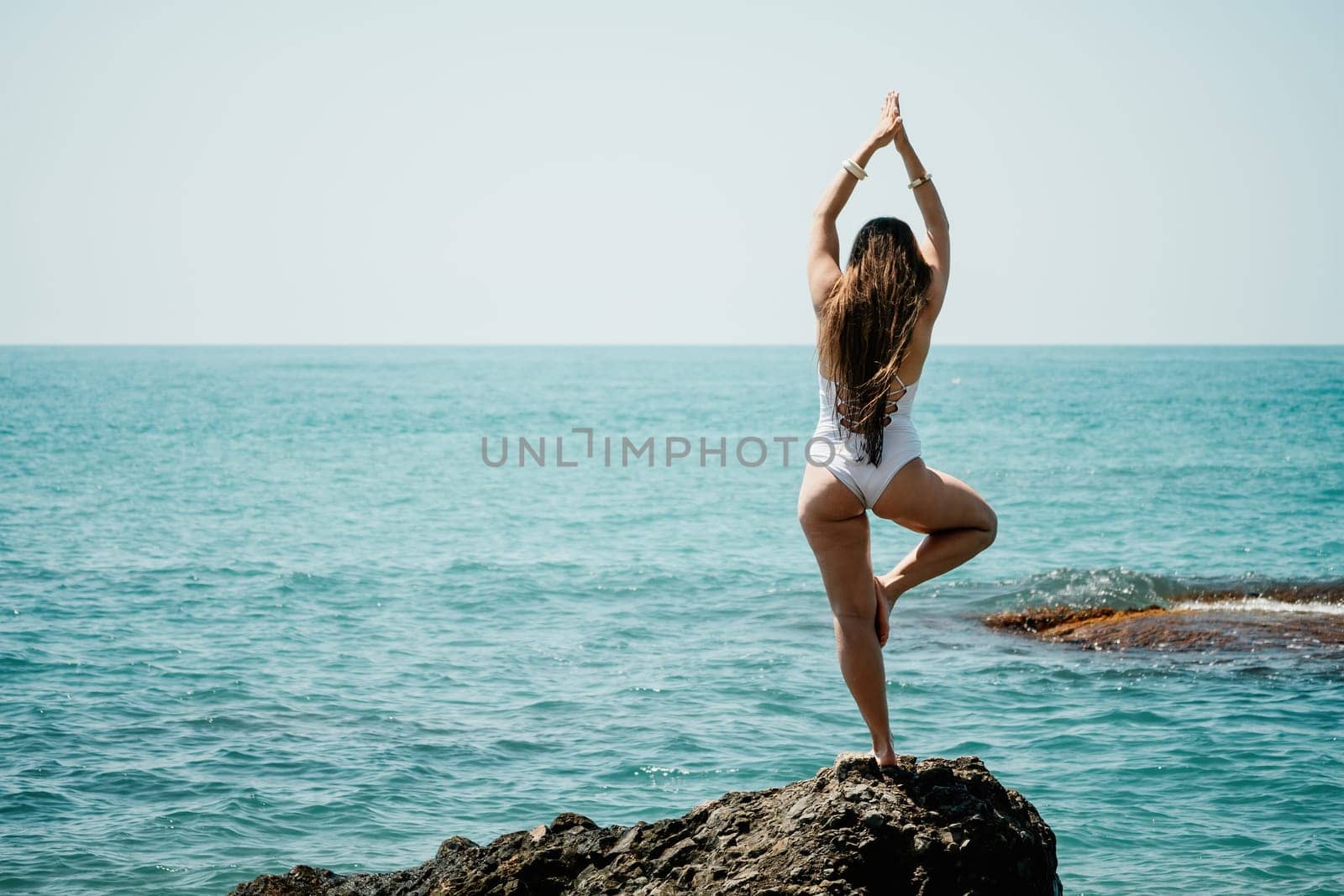 Woman sea yoga. Back view of free calm happy satisfied woman with long hair standing on top rock with yoga position against of sky by the sea. Healthy lifestyle outdoors in nature, fitness concept