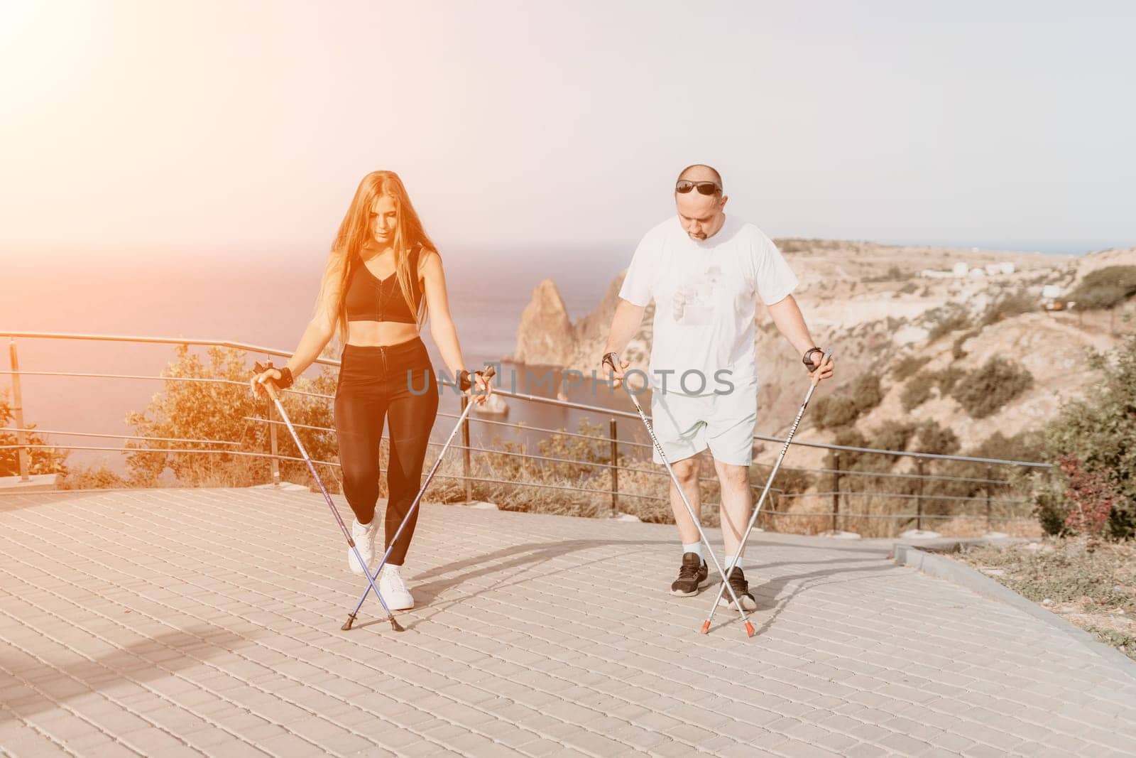 Happy Middle aged couple or friends practicing nordic walking in park near sea. Mature couple with trekking poles walking, practicing Nordic walking outdoors. Aging youthfully and sport concept.