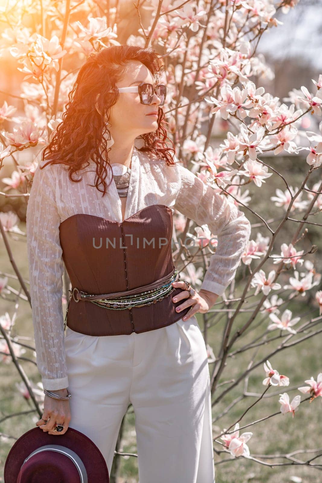 Magnolia park woman. Stylish woman in a hat stands near the magnolia bush in the park. Dressed in white corset pants and posing for the camera