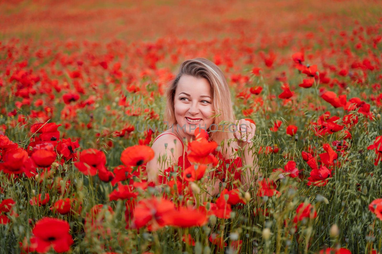 Happy woman in a red dress in a beautiful large poppy field. Blond sits in a red dress, posing on a large field of red poppies by Matiunina