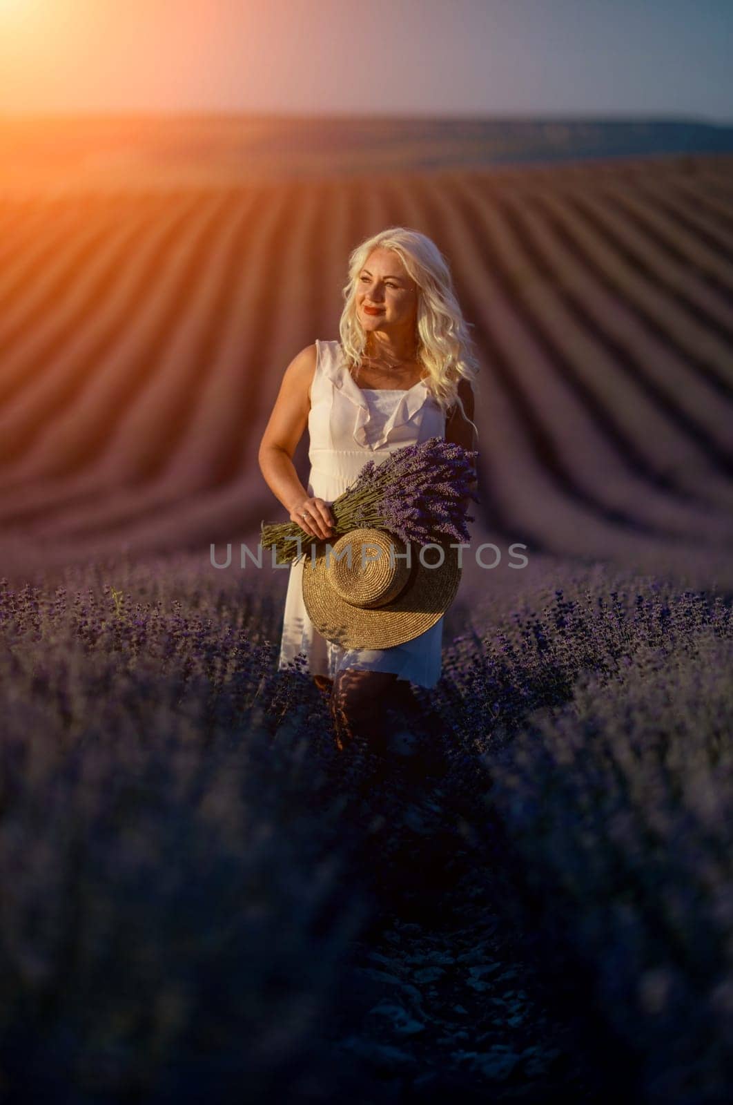 Blonde woman poses in lavender field at sunset. Happy woman in white dress holds lavender bouquet. Aromatherapy concept, lavender oil, photo session in lavender by Matiunina