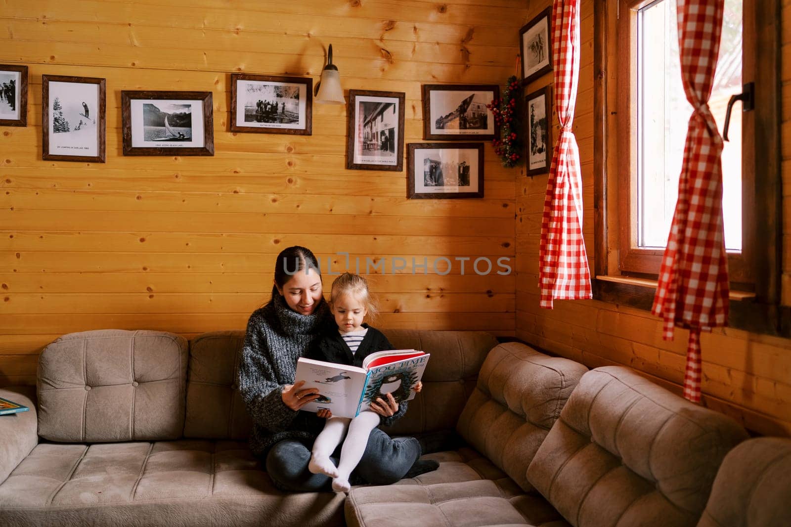 Mom reads a fairy tale to a little girl sitting on her lap on a sofa in a wooden house. High quality photo