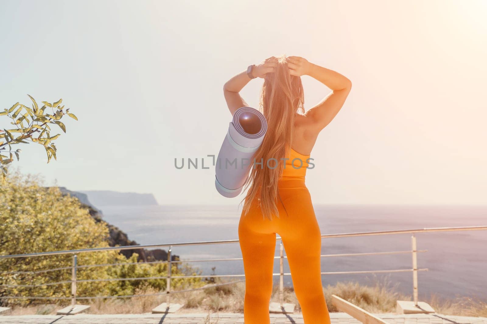 Fitness woman sea. Happy middle aged woman in orange sportswear exercises morning outdoors on yoga mat with laptop in park over ocean beach. Female fitness pilates yoga routine. Healthy lifestyle. by panophotograph