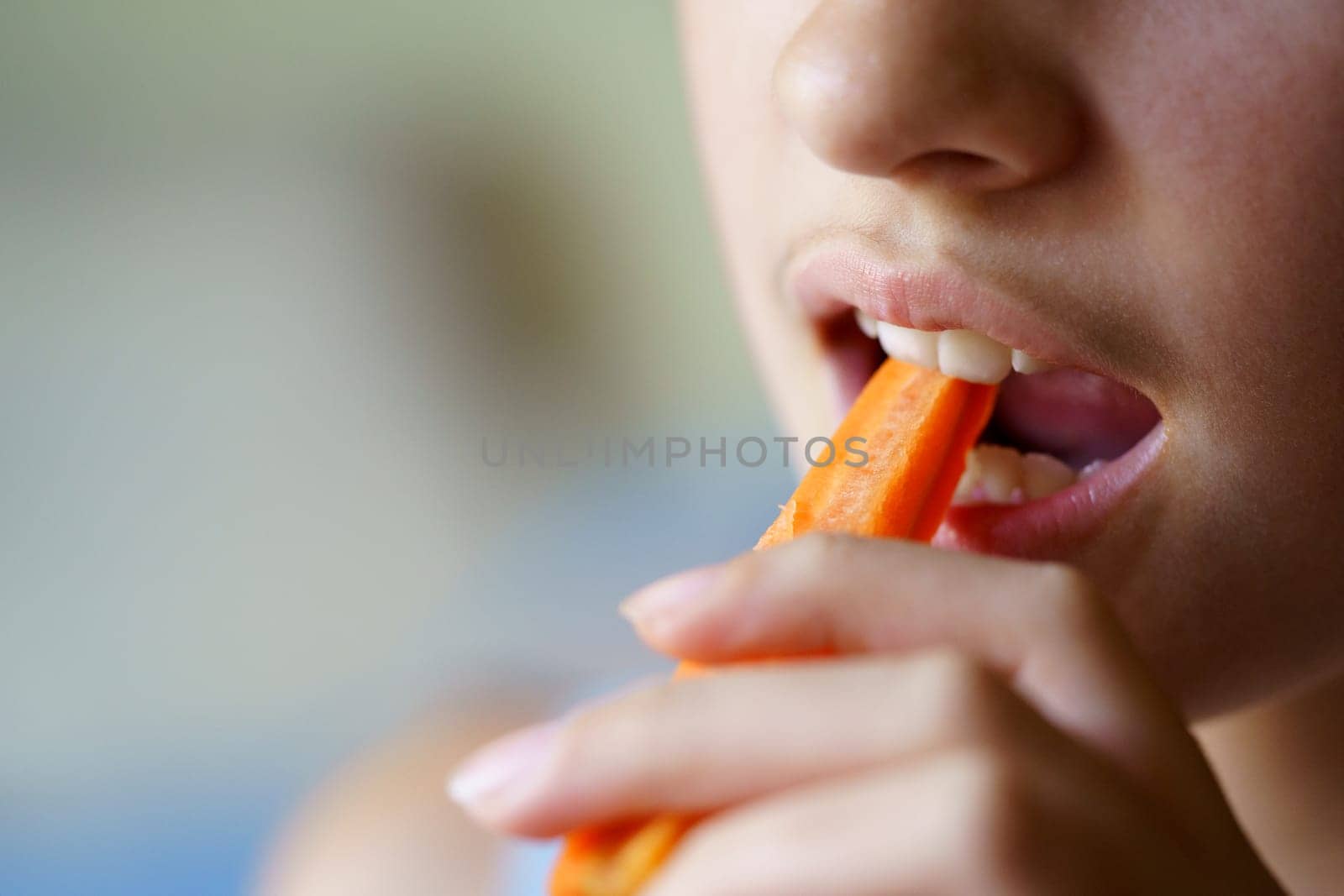 Closeup of crop anonymous teenage girl biting fresh carrot slice at home