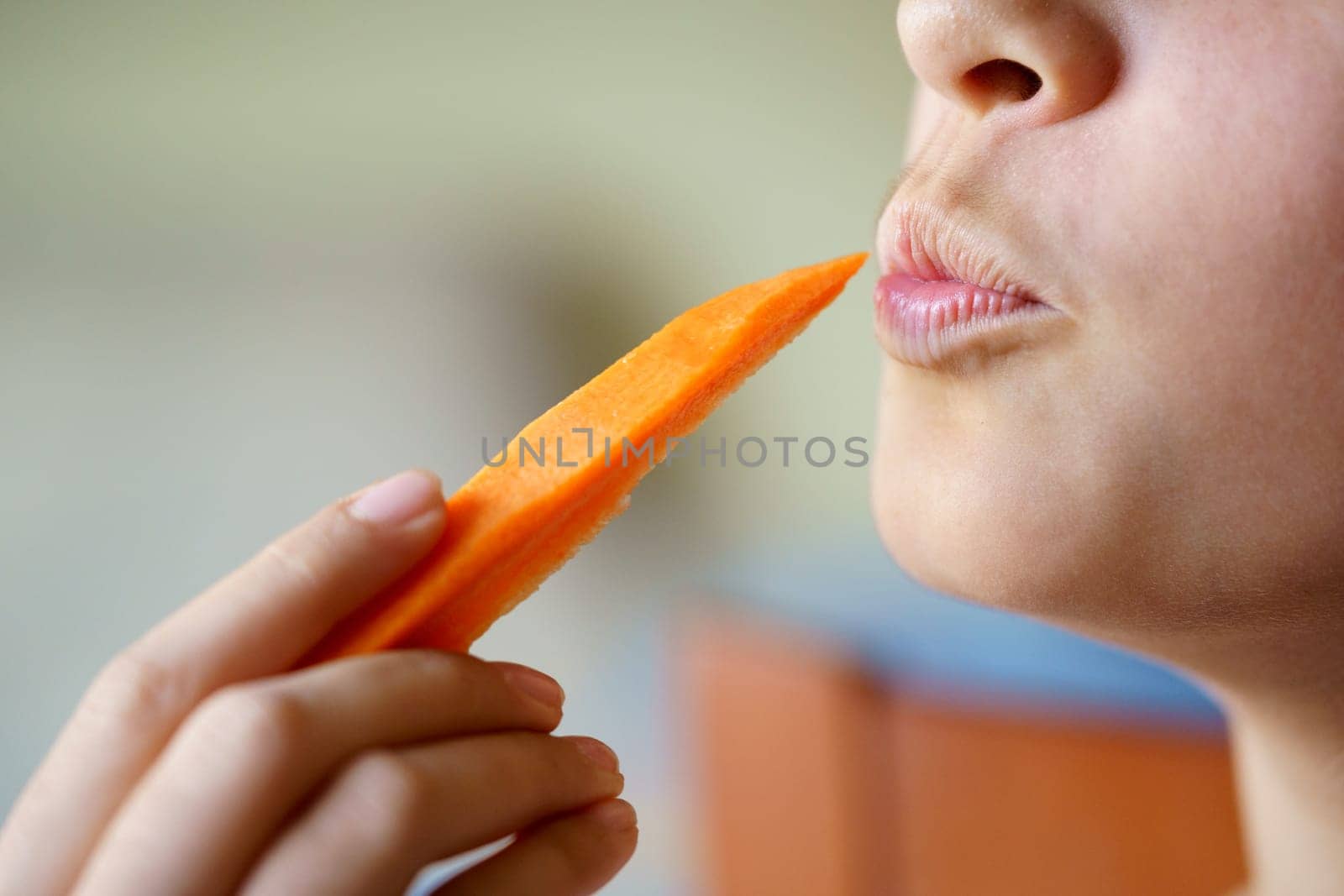 Crop anonymous young girl puckering lips while holding fresh carrot slice at home