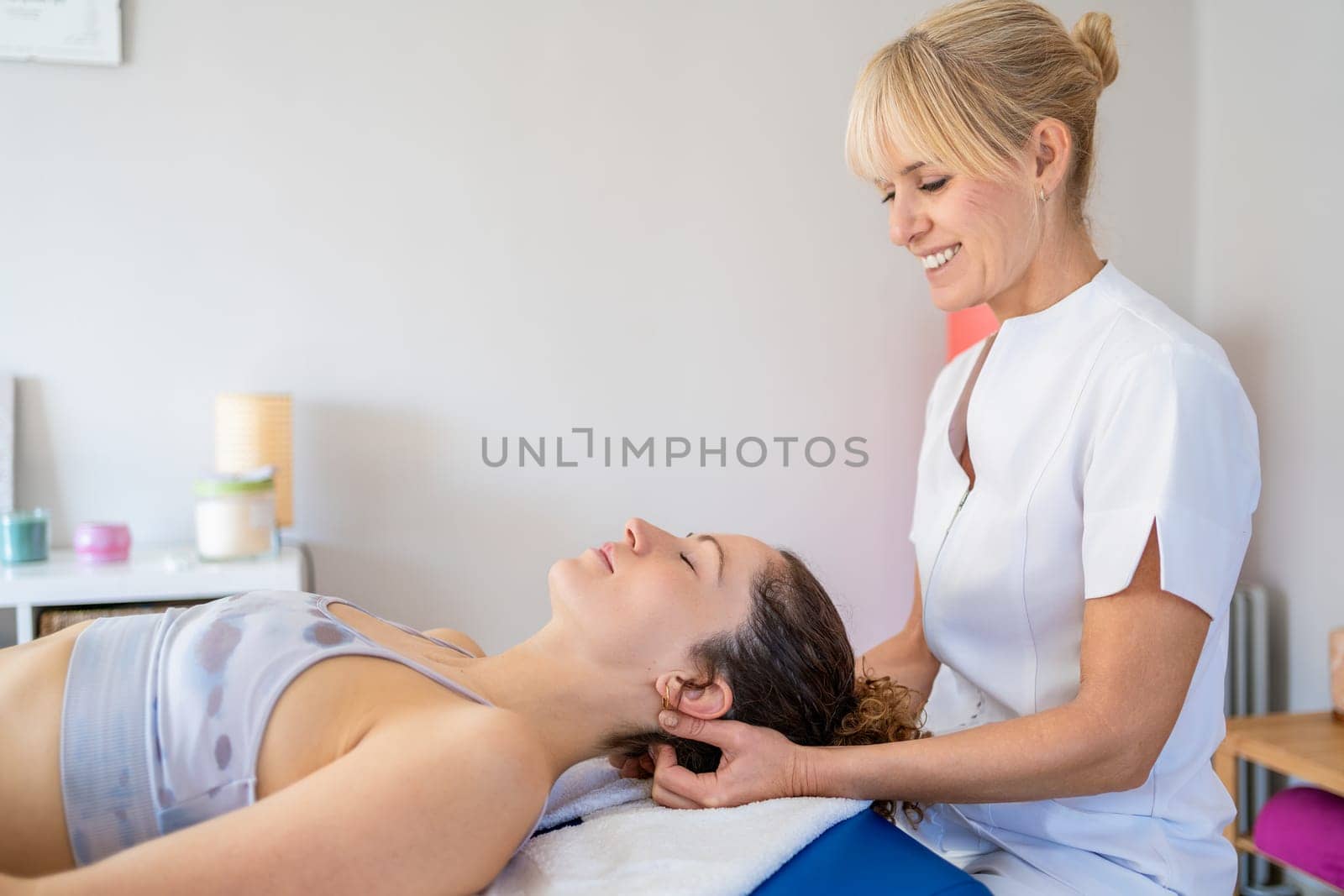 Side view of smiling female blond hair physiotherapist rubbing neck of client lying face up in bed and closing eyes during massage treatment