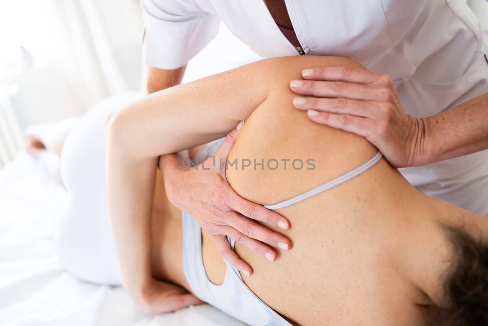 Crop anonymous female physical therapist rubbing shoulder of patient lying on couch during rehabilitation process in manual therapy center