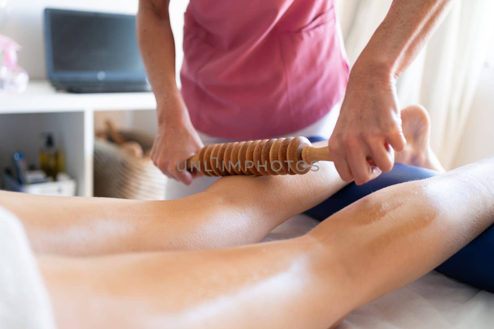 Crop anonymous female osteopathy therapist using wooden device while massaging leg of patient during physiotherapy session in bright modern clinic