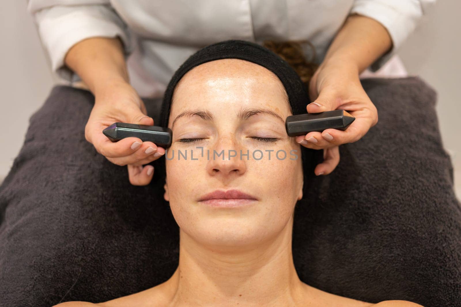 Top view of crop anonymous female cosmetician massaging face of adult woman with black crystals during skincare procedure in beauty salon