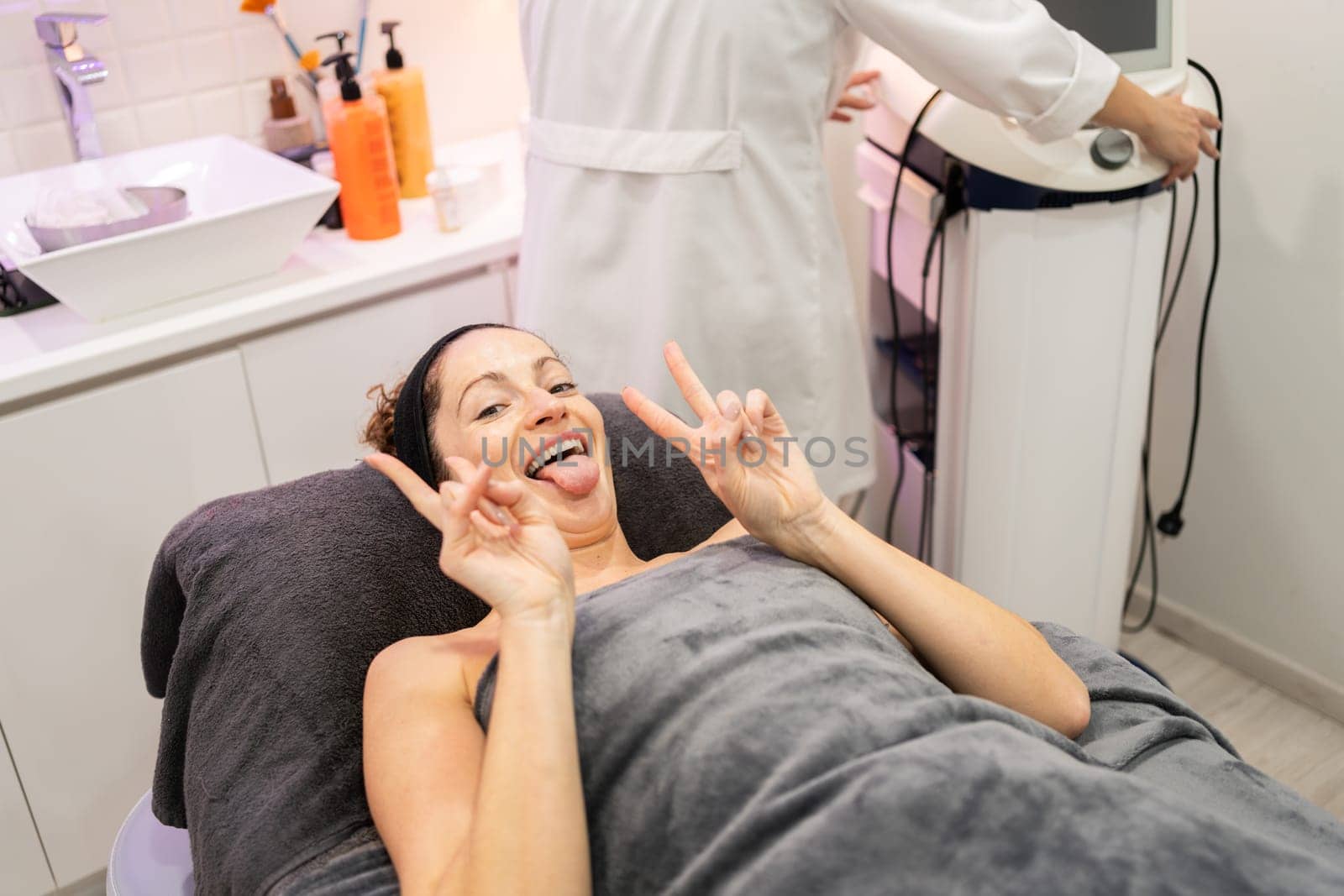 High angle of adult female with funny face and tongue out, looking at camera while lying on beauty salon bed with unrecognizable worker at equipment and showing V sign