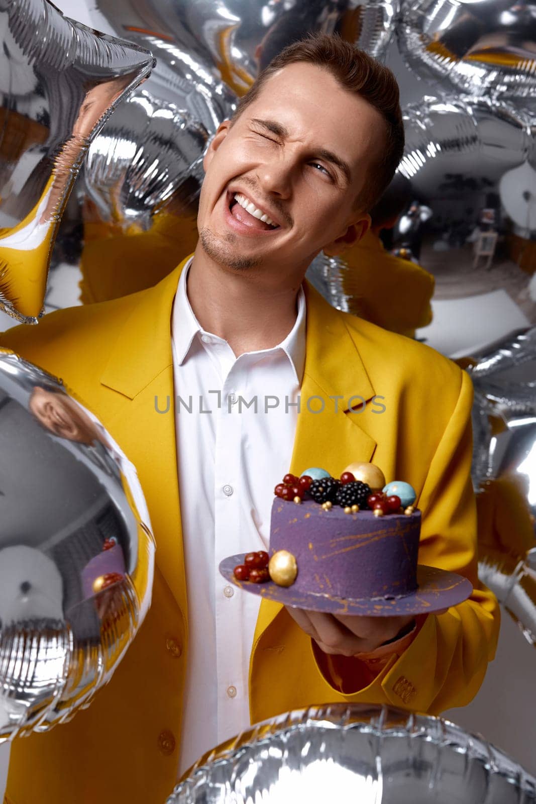 handsome birthday guy in yellow jacket holding cake looking at the camera being lovely on silver helium balloons background.