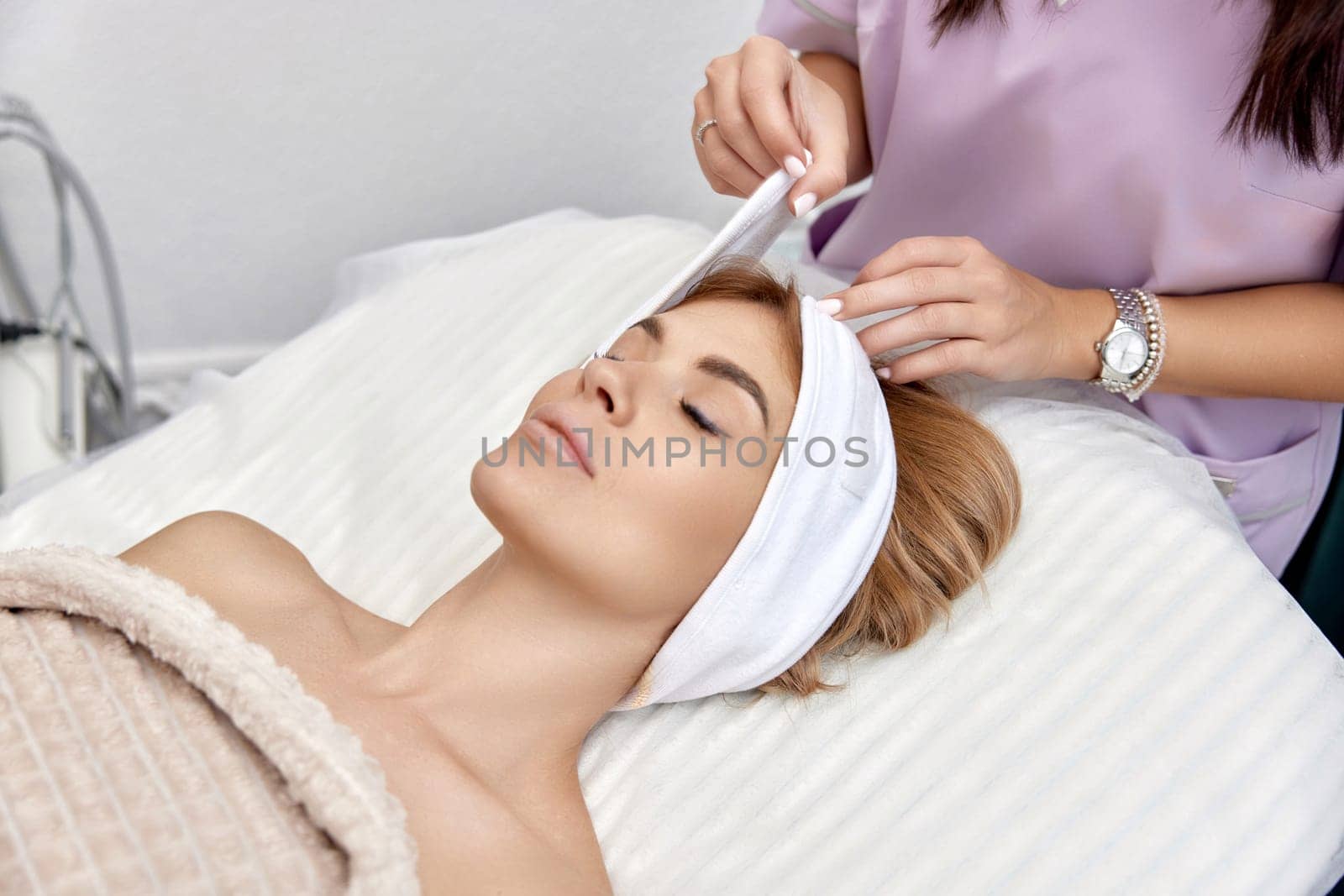 Cosmetologist putting white headband towel on female patient head in beauty salon