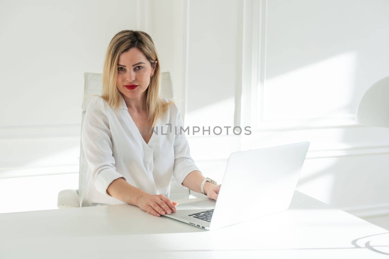 Delighted young blonde woman sitting at white table in blouse using laptop. High quality photo