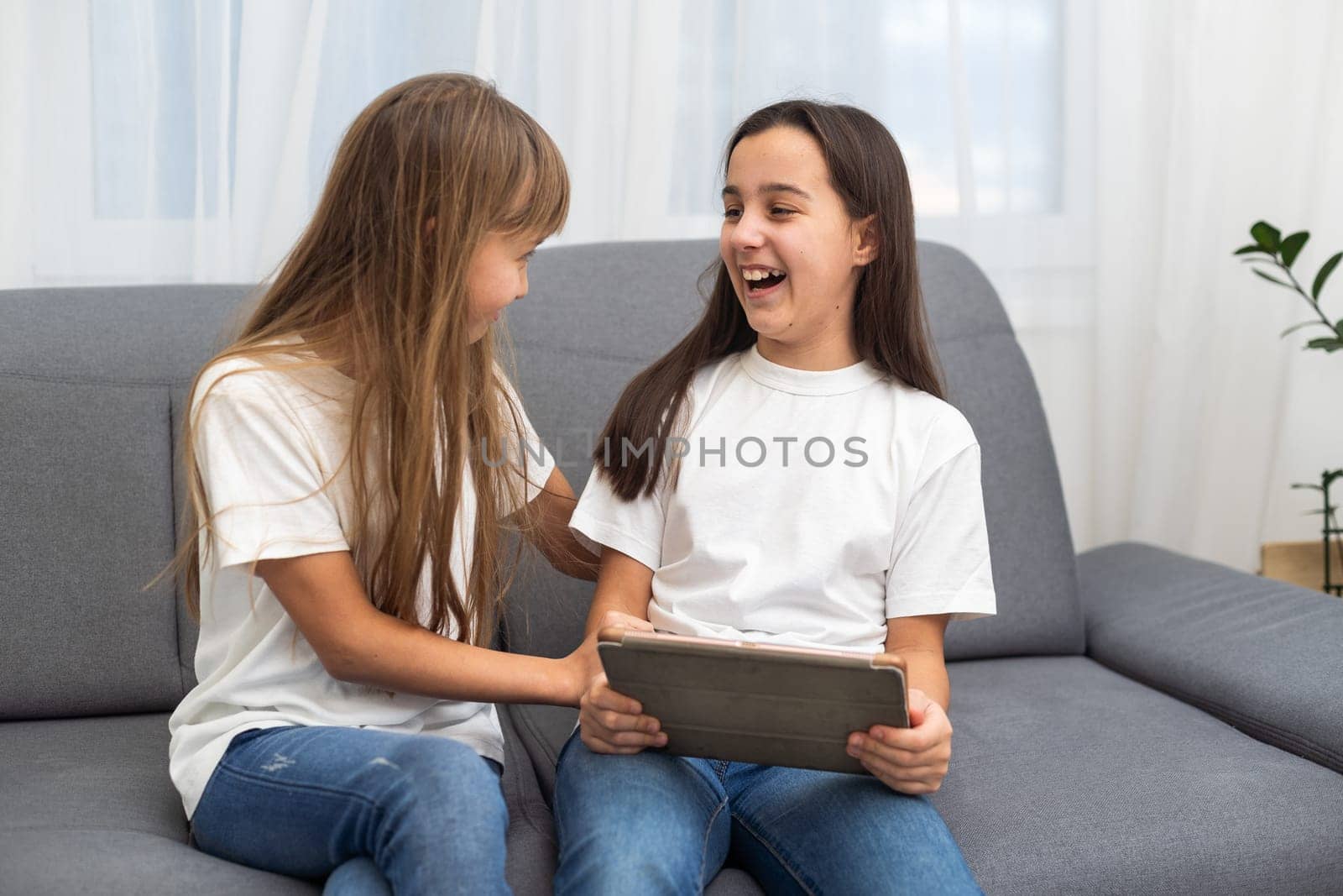 Cute little girl and older sister playing together smiling and having bonding time using a laptop on couch at home. Happy family Siblings relationship and digital technology lifestyle concept. by Andelov13