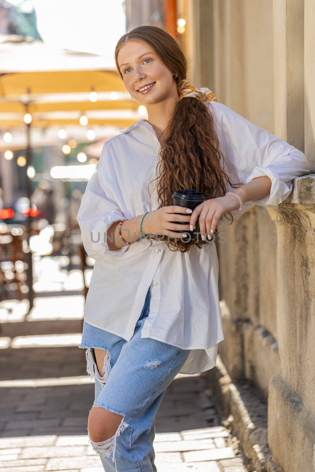 Happy redhead teenager girl enjoying morning coffee hot drink and smiling. Relaxing, taking a break. Young woman standing in urban city center street, drinking coffee to go. Town outside. Vertical