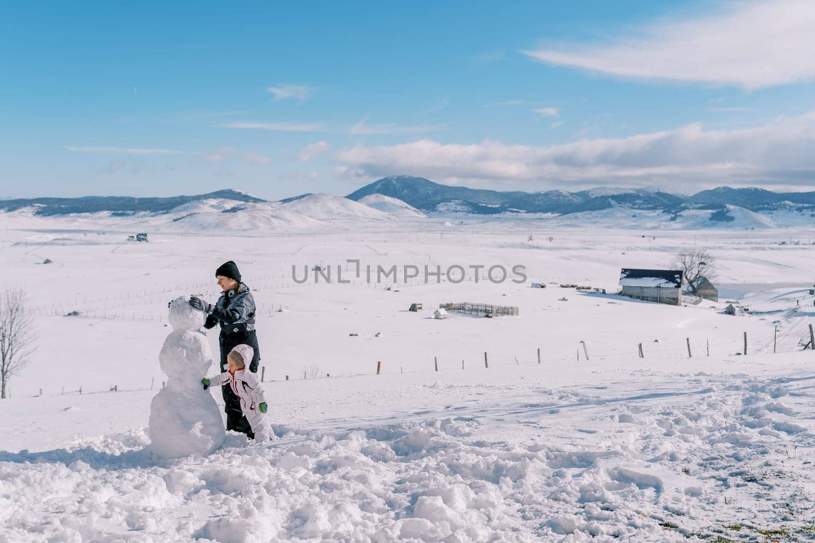Little girl helps her mother build a snowman in a mountain valley. Side view. High quality photo