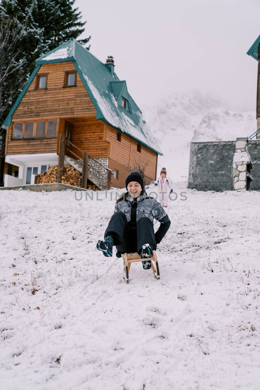 Little girl stands near the house and looks at her laughing mother riding down the hill on a sled. High quality photo