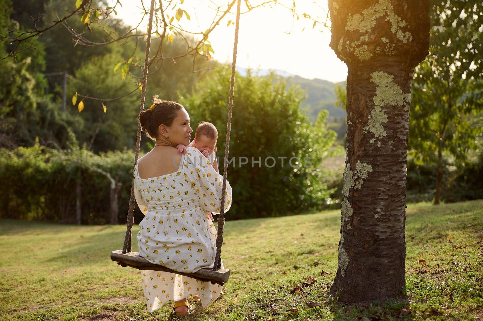 Rear view of a young adult mother holding her newborn baby while sitting on the swing in the park at sunset by artgf