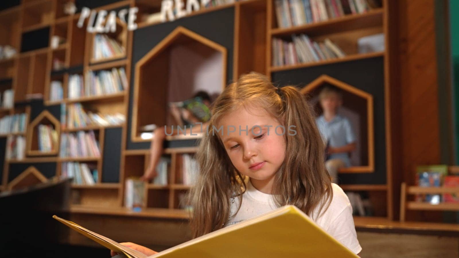 Young smart caucasian girl picking reading a book while sitting at library. Clever child learning, studying, open a books at library. Attractive kid turning page with blurring background. Erudition.