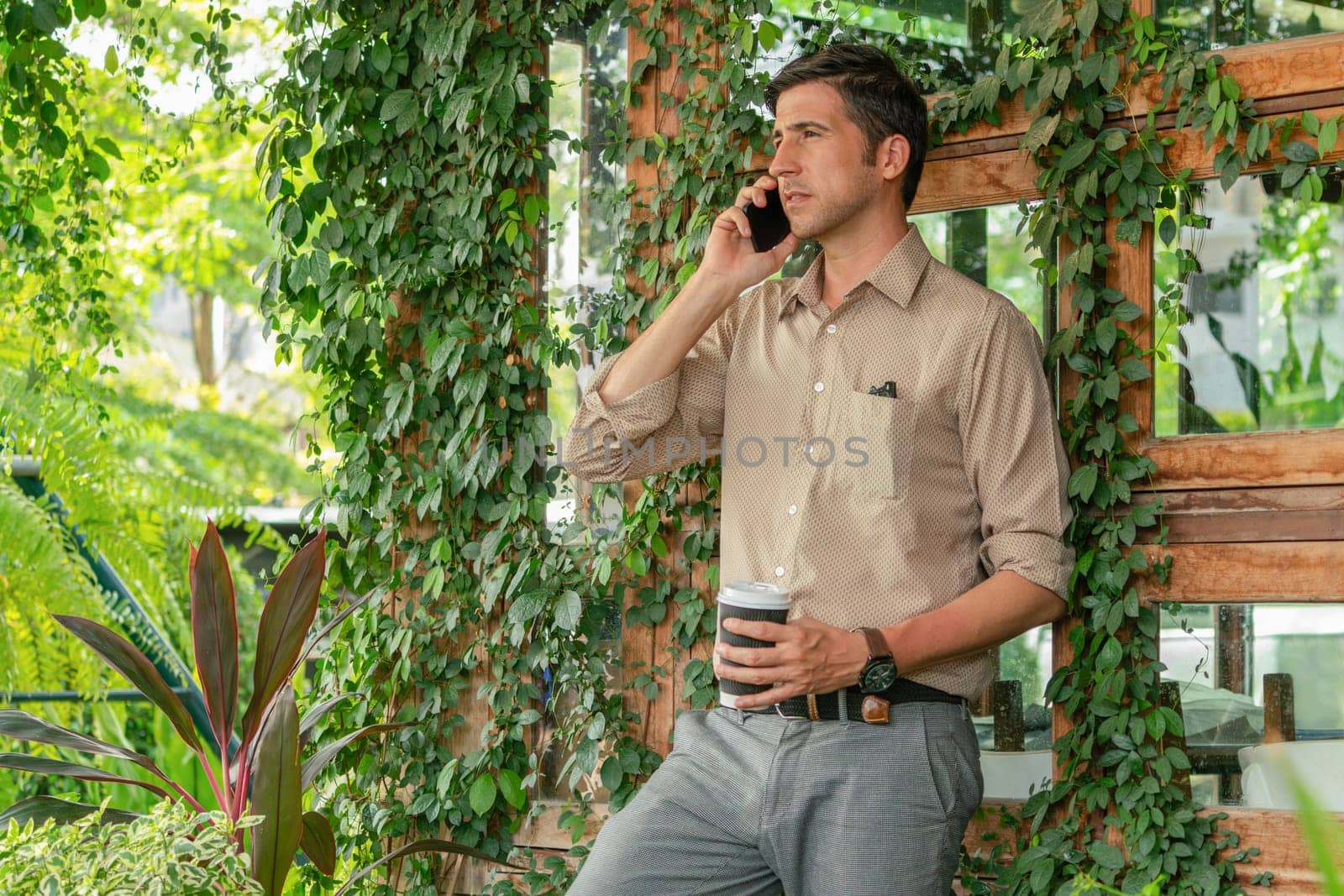 Young handsome man walk out of coffee shop with cafe garden, holding a coffee cup and a shopping bag while talking on the phone. Modern happy carefree with sunglasses lifestyle. Expedient