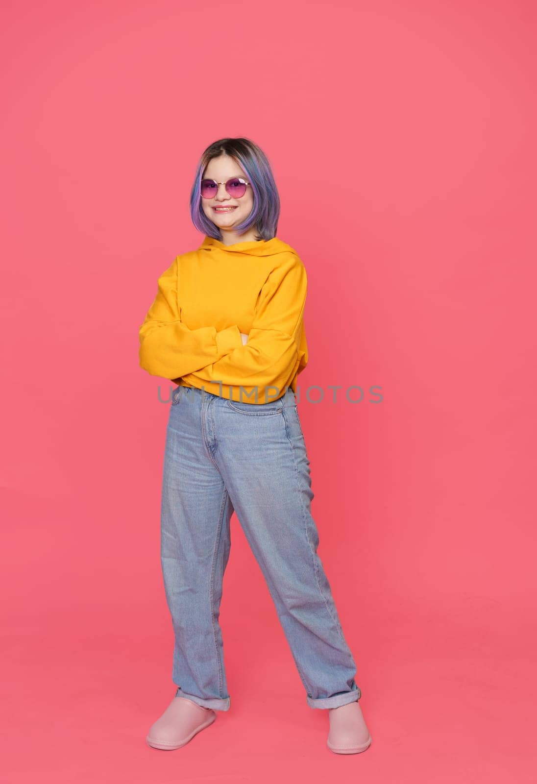Smiling Asian teenager in crossed hands position with colored hair stylishly dressed in yellow jacket, standing on pink background. Youth culture, girl's expressive style and personality in contemporary and modern setting. Full-length portrait, . High quality photo