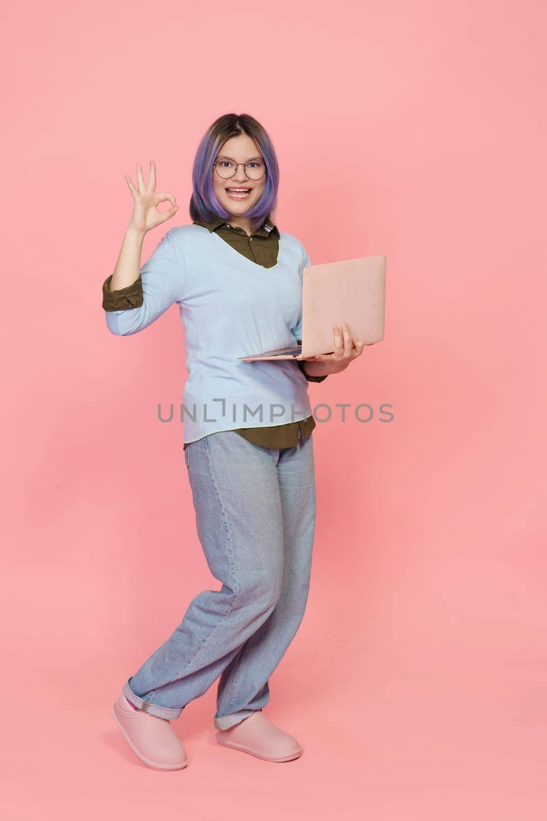 Successful student, holding opened laptop, presents confident full-length portrait, flashing OK sign. Girl in casual wear against pink background. High quality photo