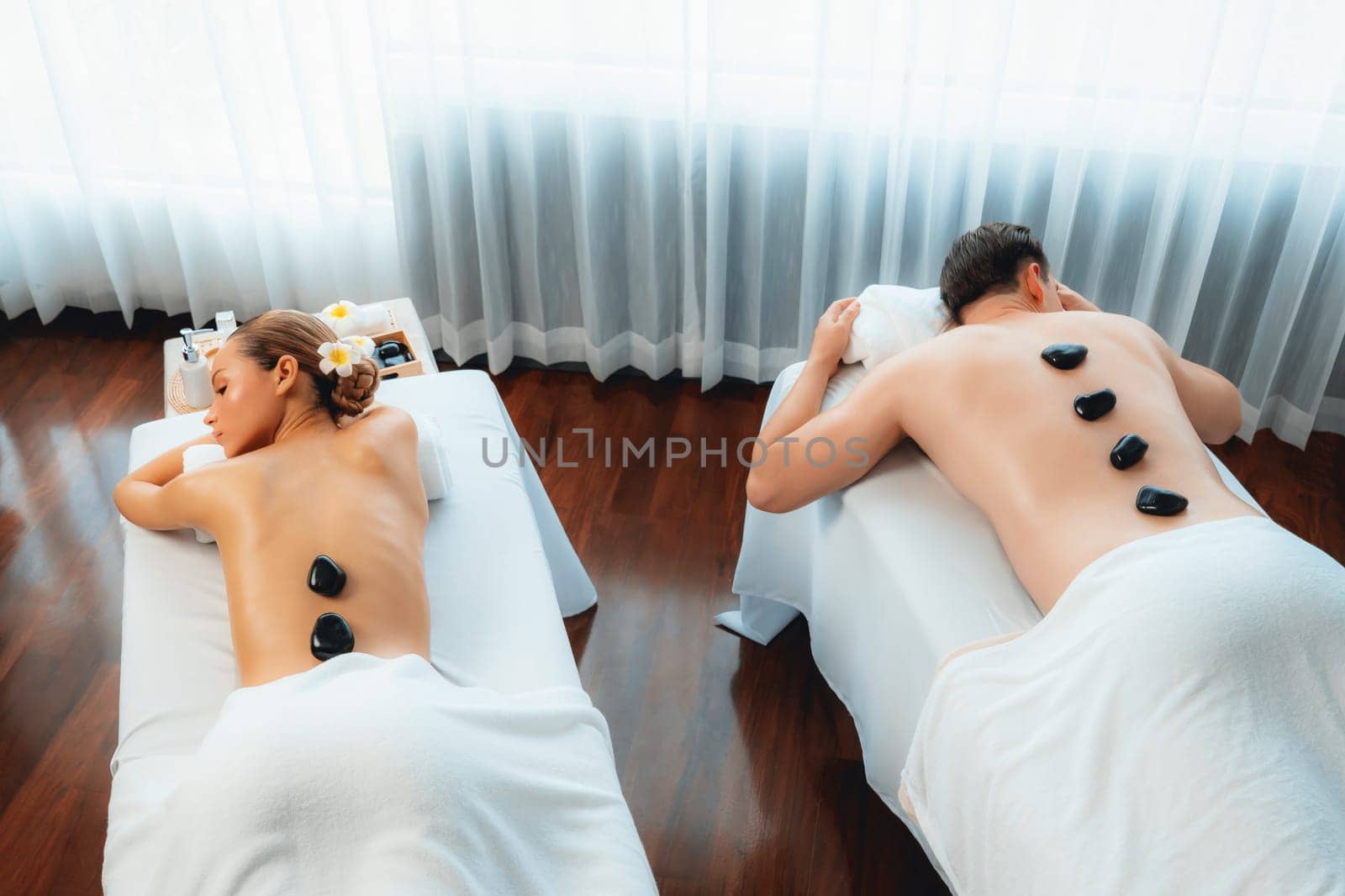 Hot stone massage at spa salon in luxury resort with day light serenity ambient, blissful couple customer enjoying spa basalt stone massage glide over body with soothing warmth. Quiescent