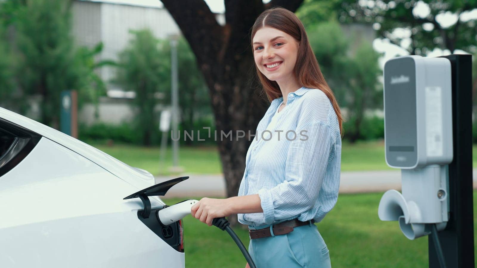 Young woman recharging battery for electric car during road trip travel EV car in natural forest or national park. Eco friendly travel during vacation and holiday. Exalt