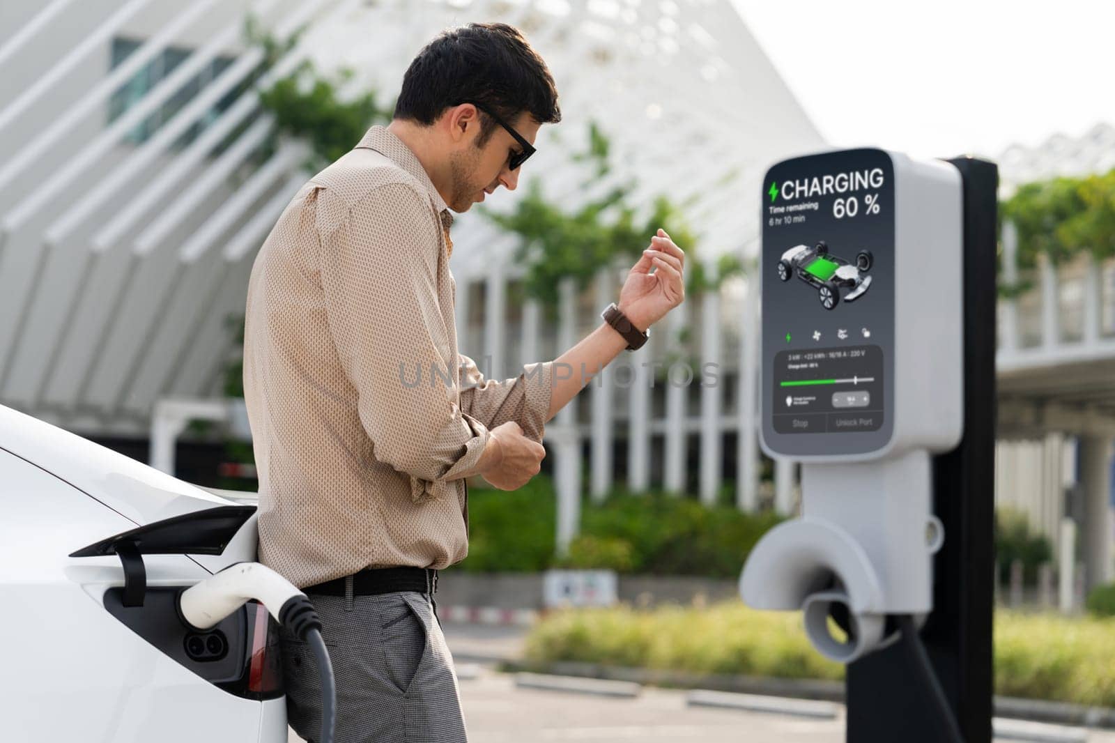 Young man put EV charger to recharge electric car's battery from charging station in city commercial parking lot. Rechargeable EV car for sustainable environmental friendly urban travel. Expedient