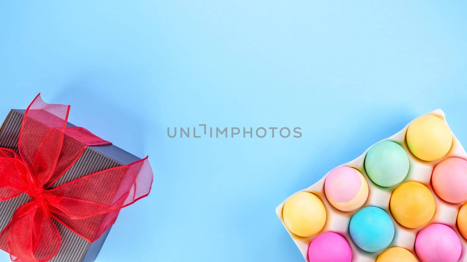 Easter pastel background with colorful eggs and a gift box with red bow on a blue background. The concept of a Happy Easter. Top view, copy space